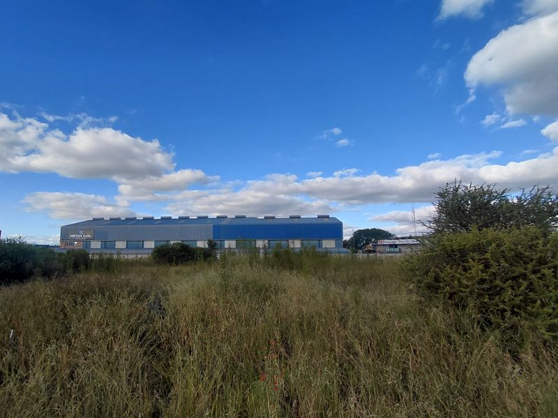 A wide-angle view of a vacant commercial land plot featuring overgrown vegetation with a large industrial warehouse building visible in the background.