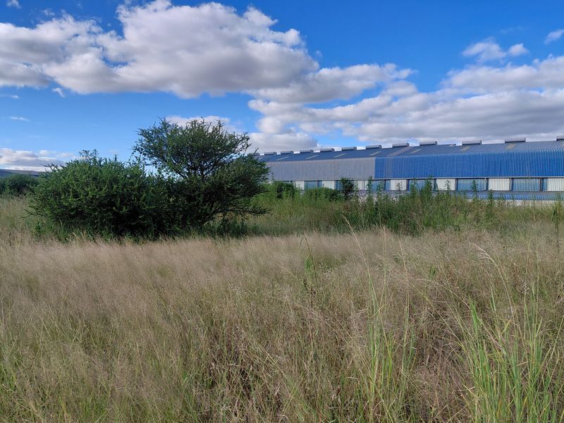 A view of a vacant commercial land plot featuring overgrown grass and shrubs with a large industrial warehouse building visible in the background.