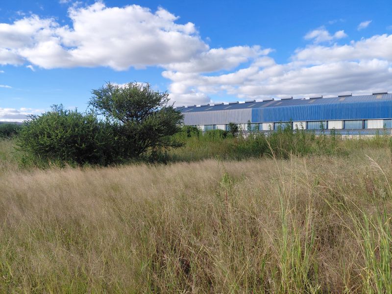A view of a vacant commercial land plot featuring overgrown grass and shrubs, with a large industrial warehouse building visible in the background.