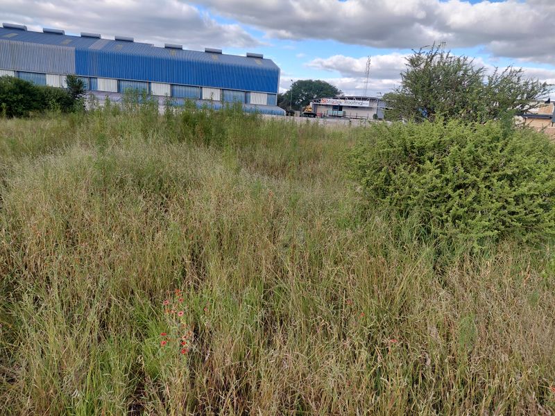 A view of a vacant commercial land plot featuring overgrown grass and shrubs, situated in an industrial area.