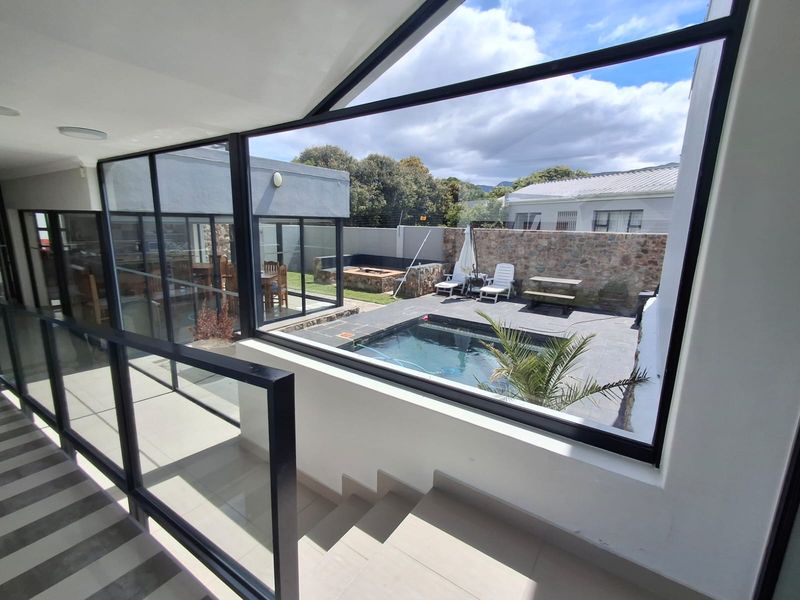 A bright hallway featuring a modern glass balustrade and large windows overlooking a private courtyard with a swimming pool and stone wall.