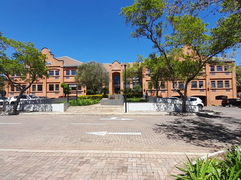 Front elevation of a multi-story brick office building featuring a paved entrance area, landscaped gardens, and established trees.