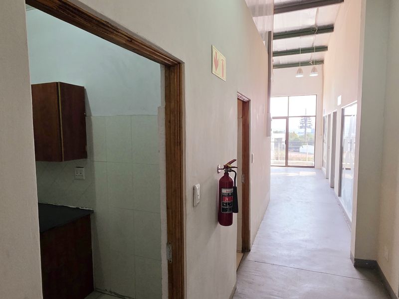 A bright industrial hallway featuring polished concrete-style flooring, exposed ceiling beams, and glass-partitioned office spaces with a wall-mounted fire extinguisher.