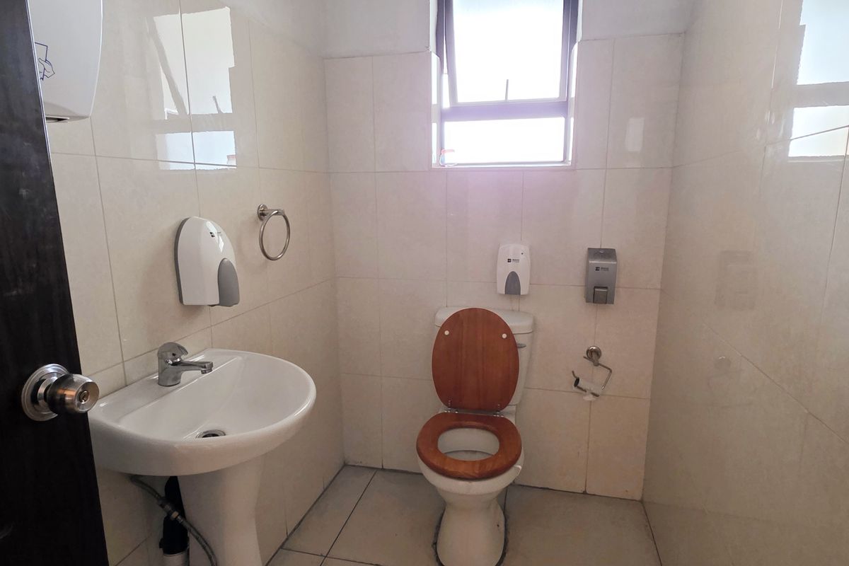 A functional industrial-style bathroom featuring a pedestal washbasin, toilet, and full-height ceramic wall tiling with a small casement window for ventilation.