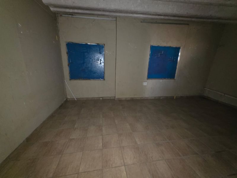 An empty commercial interior space featuring tiled flooring, exposed concrete ceiling, and two windows secured with blue metal shutters.