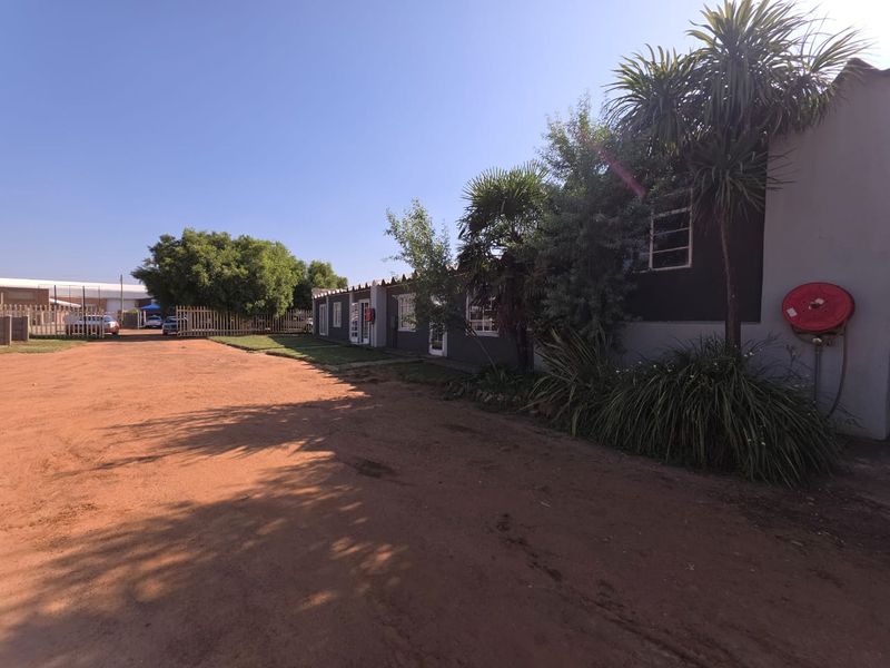 Exterior view of a commercial property featuring a long building facade, mature landscaping, and an unpaved access driveway.