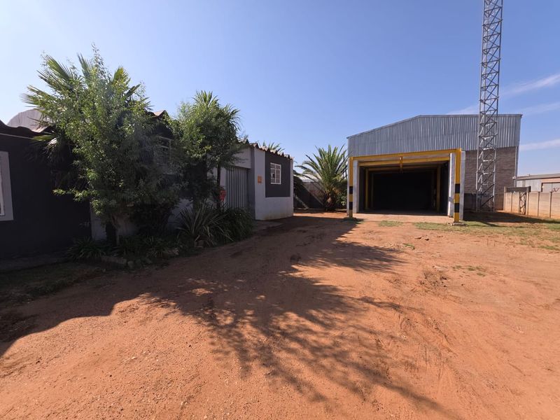 Commercial property exterior featuring a large warehouse structure with a high-clearance entrance and an adjacent office or utility building on a dirt lot.