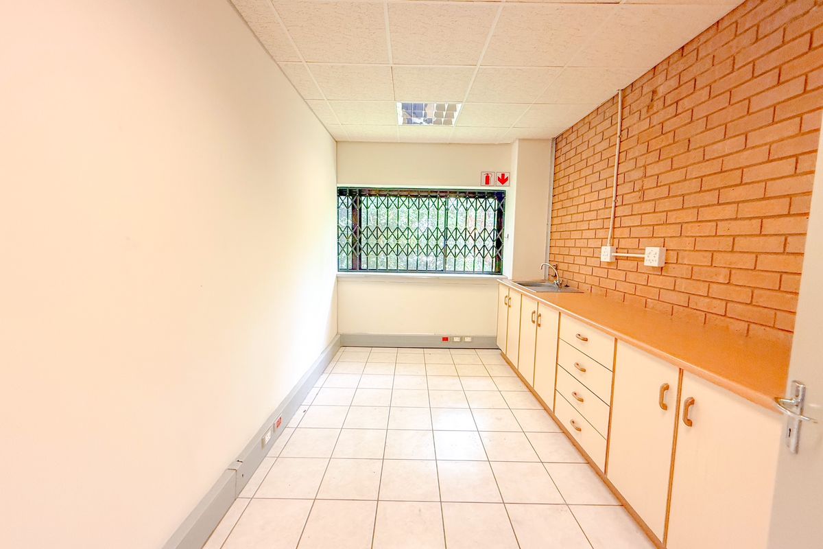 A functional office kitchenette featuring built-in cabinetry, a stainless steel sink, tiled flooring, and a security gate on the window.
