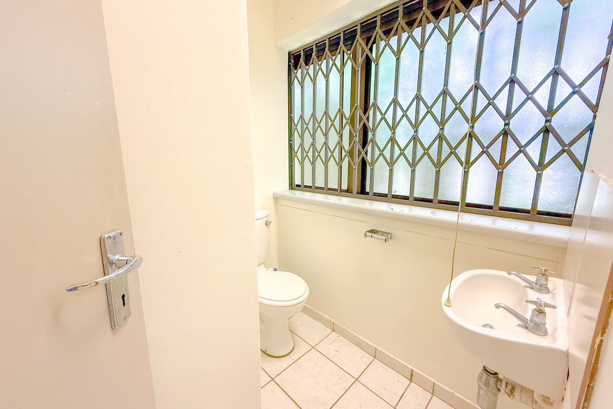A functional office restroom featuring a white ceramic toilet, wall-mounted basin, and a window secured with a metal security gate.