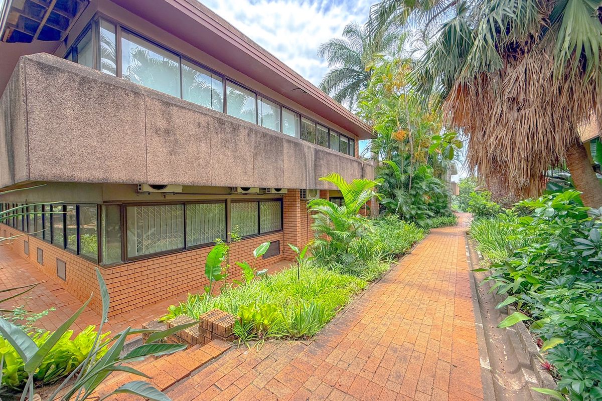 Exterior view of a multi-level office building featuring brick and concrete construction, large windows, and a paved walkway surrounded by established garden landscaping.