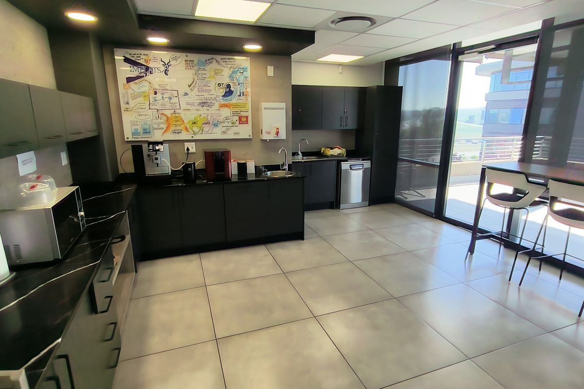 Modern office kitchen featuring sleek dark cabinetry, integrated sink, dishwasher, and large sliding glass doors providing ample natural light and access to the balcony.