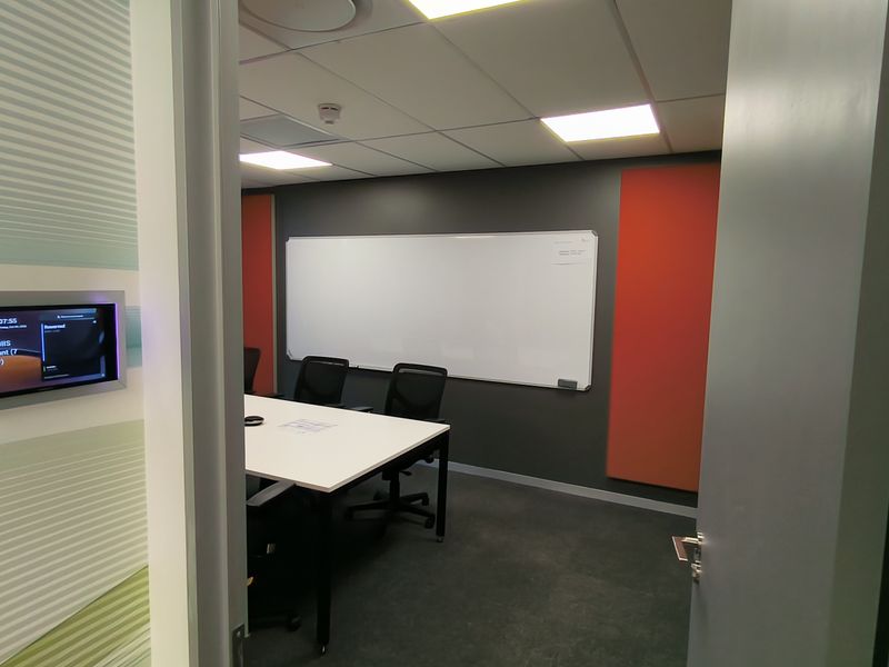 A modern, well-maintained office meeting room featuring acoustic wall panels, a large whiteboard, recessed ceiling lighting, and an integrated room booking system.