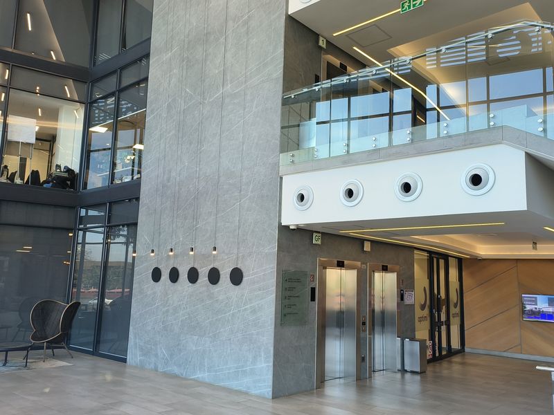 Modern office building lobby featuring a double-volume glass facade, stone-clad feature wall, stainless steel elevators, and a contemporary glass-railed mezzanine level.