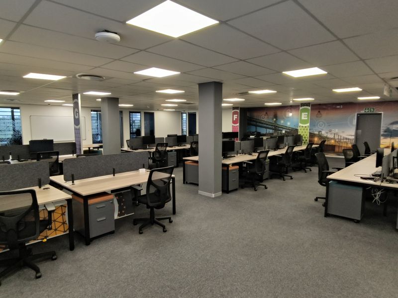 A spacious open-plan office area featuring modern workstations, carpeted flooring, suspended ceilings with recessed lighting, and integrated structural columns.