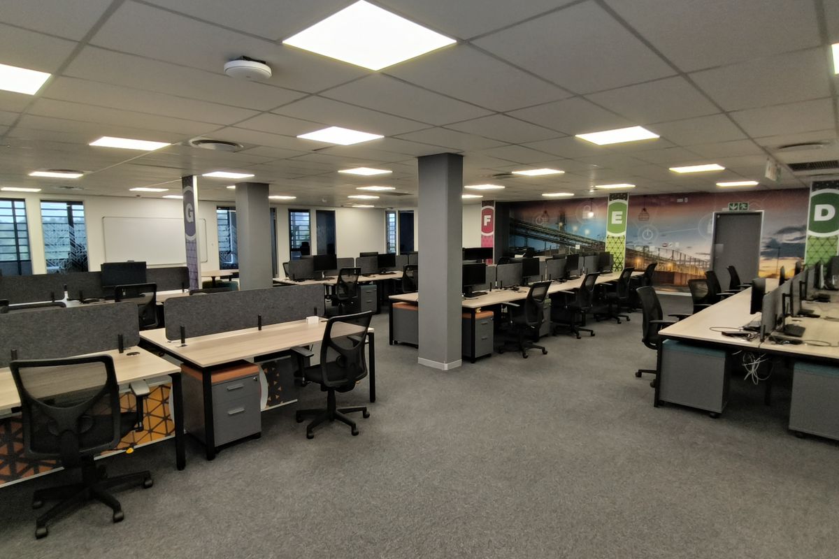 A spacious open-plan office area featuring modern workstations, carpeted flooring, suspended ceilings with recessed lighting, and integrated structural columns.