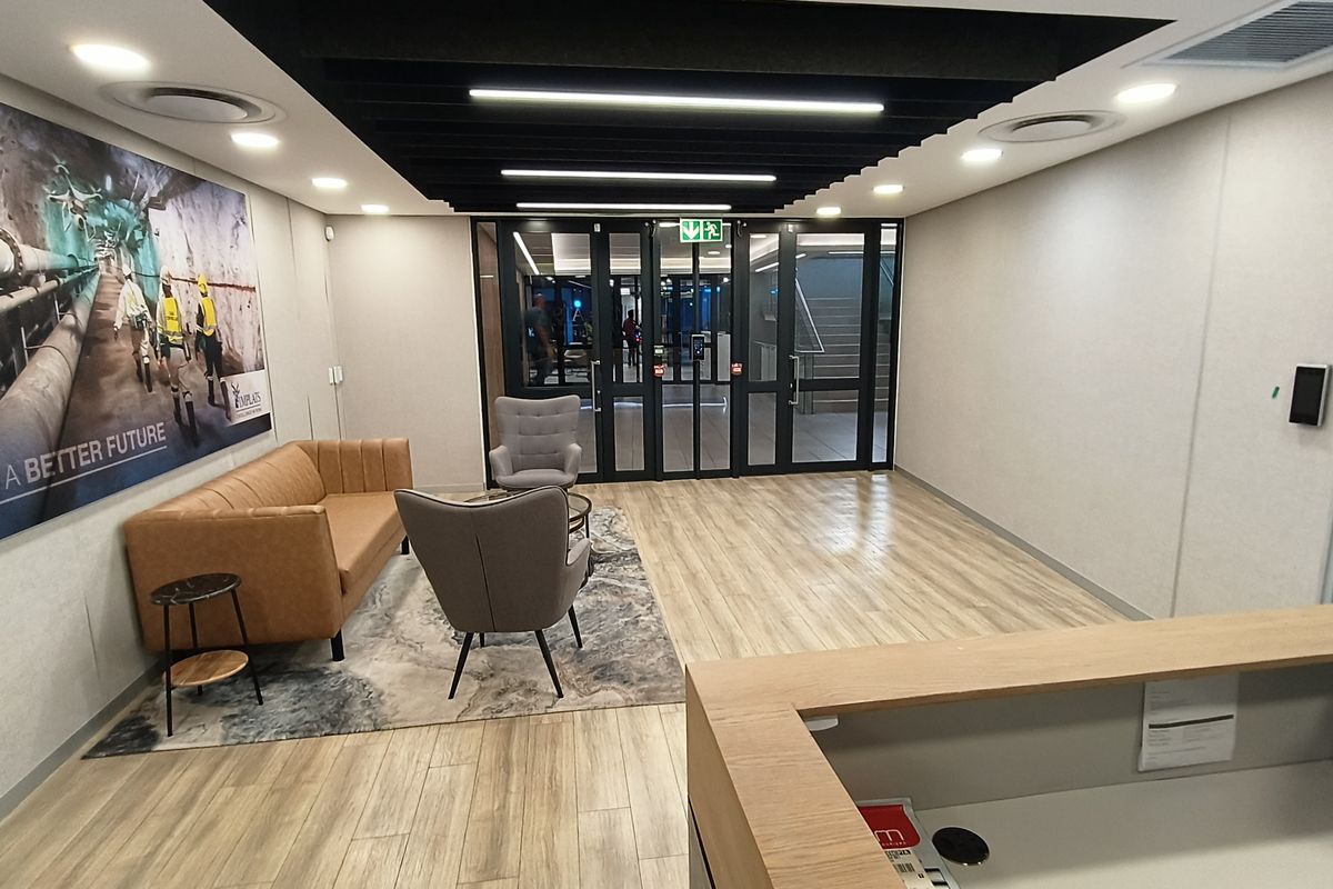Modern office reception area featuring wood-look flooring, recessed lighting, black acoustic ceiling baffles, and a professional reception desk with glass entry doors.