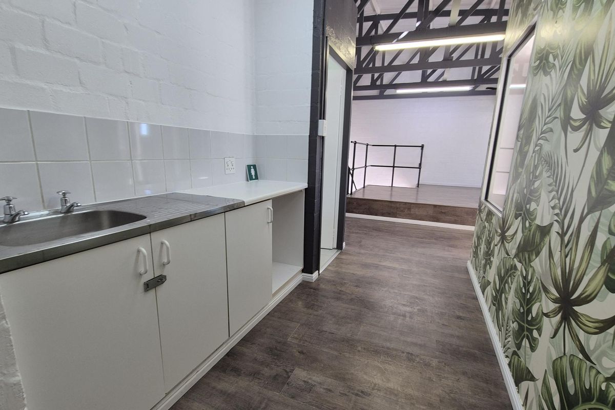 A functional kitchenette area featuring built-in cabinetry, a stainless steel sink, and wood-look flooring, leading into an open-plan office space with exposed roof trusses.