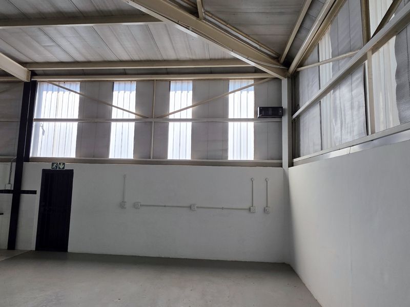 Spacious industrial utility area featuring high ceilings with exposed steel trusses, translucent natural lighting panels, and a functional kitchenette corner with built-in cabinetry.