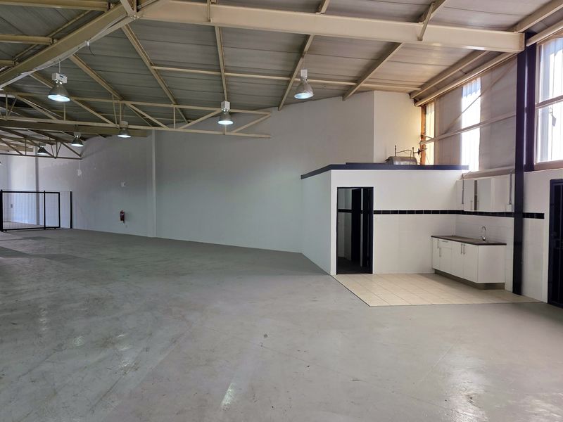 Spacious industrial warehouse unit featuring high ceilings with exposed steel trusses, concrete flooring, a small kitchenette area, and secure access points.