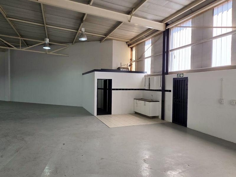 Spacious industrial warehouse unit featuring high ceilings with exposed steel trusses, concrete flooring, and a dedicated kitchenette area with sink and cabinetry.