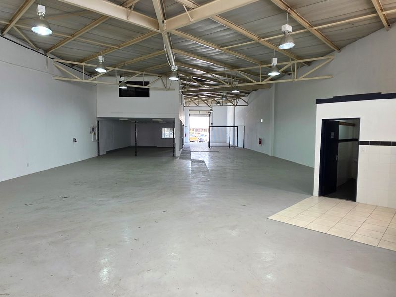Spacious industrial warehouse unit featuring high ceilings with exposed steel trusses, concrete flooring, a mezzanine level, and a functional kitchenette area.