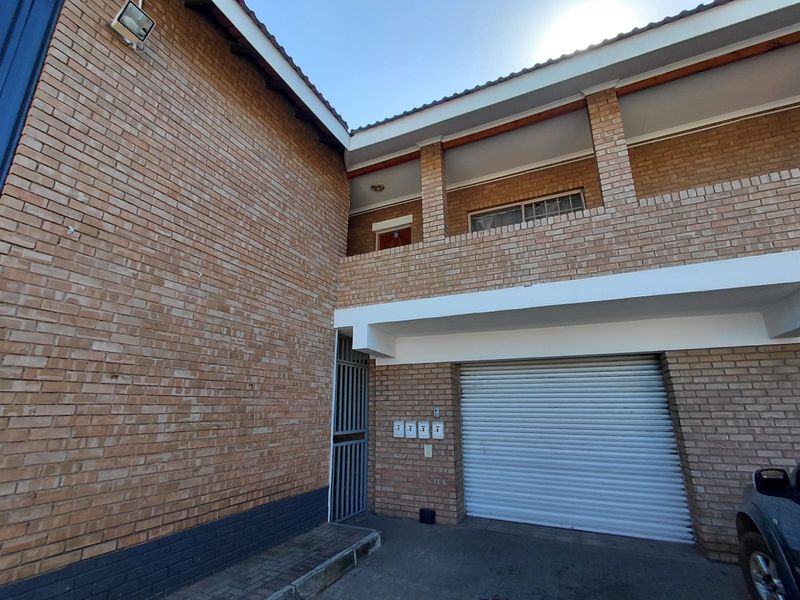 Exterior view of a multi-level brick building featuring a secure roller shutter garage door, covered balcony, and paved entrance area.