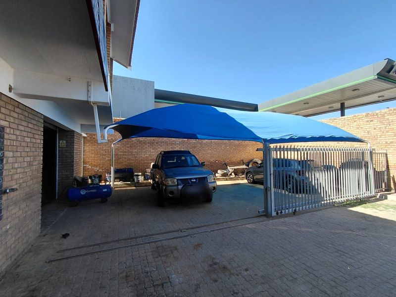Outdoor covered parking area featuring a blue shade sail structure, brick walling, and paved flooring, suitable for business vehicle storage.