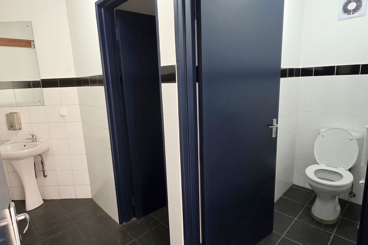 A clean, functional industrial-style bathroom featuring a pedestal basin, toilet, and tiled finishes with dark blue door accents.
