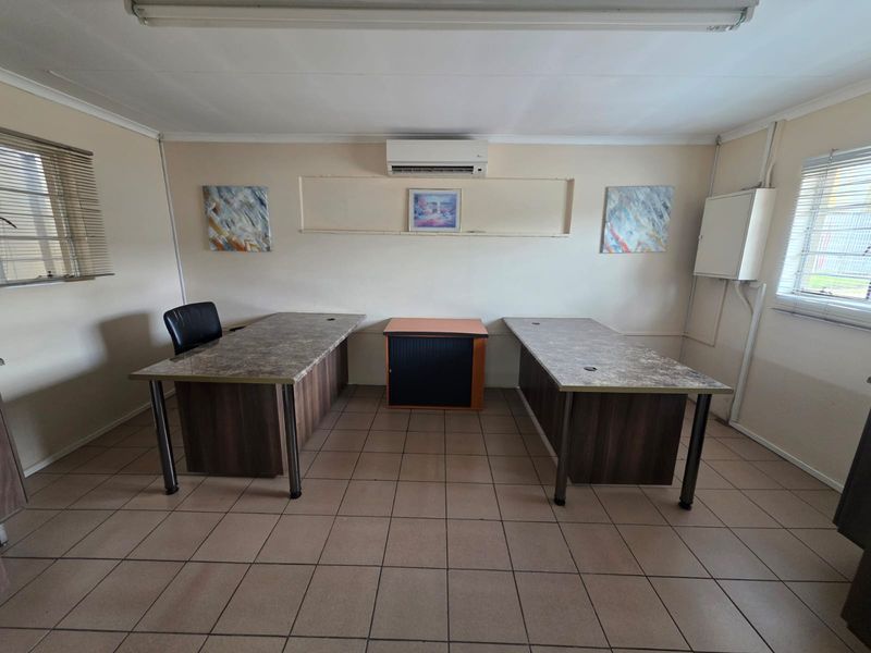 A functional office space featuring tiled flooring, a wall-mounted air conditioning unit, and dual windows with blinds.