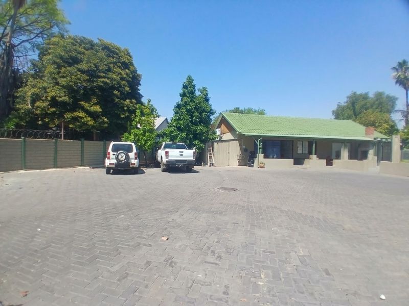 Exterior view of a commercial property featuring a large paved parking area, perimeter wall with security fencing, and a building with a tiled roof.