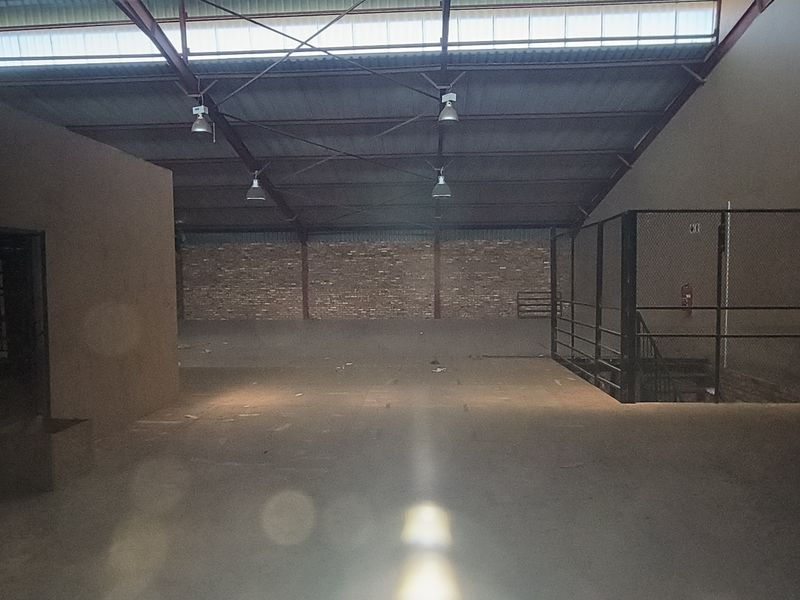 Spacious industrial warehouse interior featuring exposed steel roof trusses, high-bay lighting, concrete flooring, and a secure metal mesh enclosure.