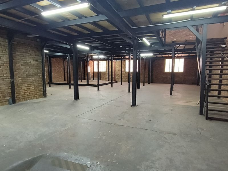 Spacious industrial open-plan area featuring exposed brick walls, a steel mezzanine structure, a staircase, and durable concrete flooring with overhead lighting.