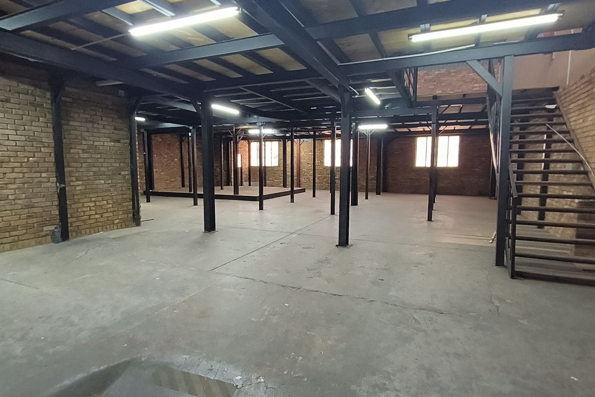Spacious industrial open-plan area featuring exposed brick walls, a steel mezzanine structure, a staircase, and durable concrete flooring with overhead lighting.