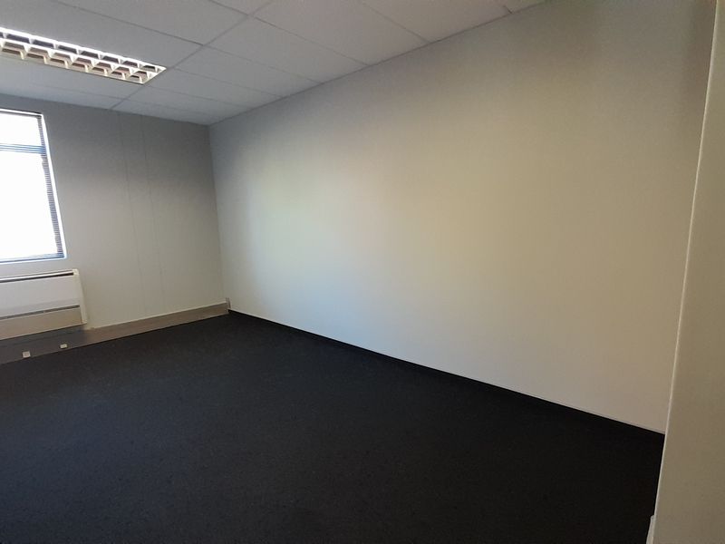 A clean, empty office space featuring dark carpeting, a suspended ceiling with recessed lighting, and a wall-mounted HVAC unit.