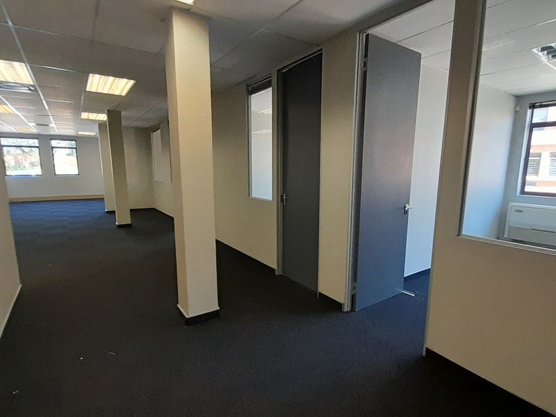 An open-plan office space featuring carpeted flooring, structural columns, suspended ceilings with recessed lighting, and glass-partitioned private office rooms.
