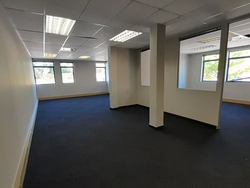An open-plan office space featuring dark carpeting, a suspended ceiling with recessed lighting, and a glass-partitioned private office area.