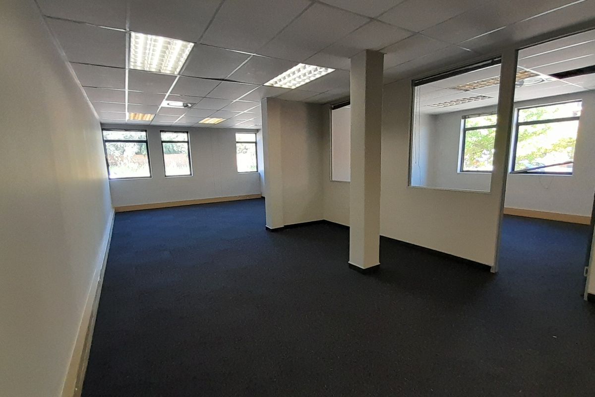 An open-plan office space featuring dark carpeting, a suspended ceiling with recessed lighting, and a glass-partitioned private office area.