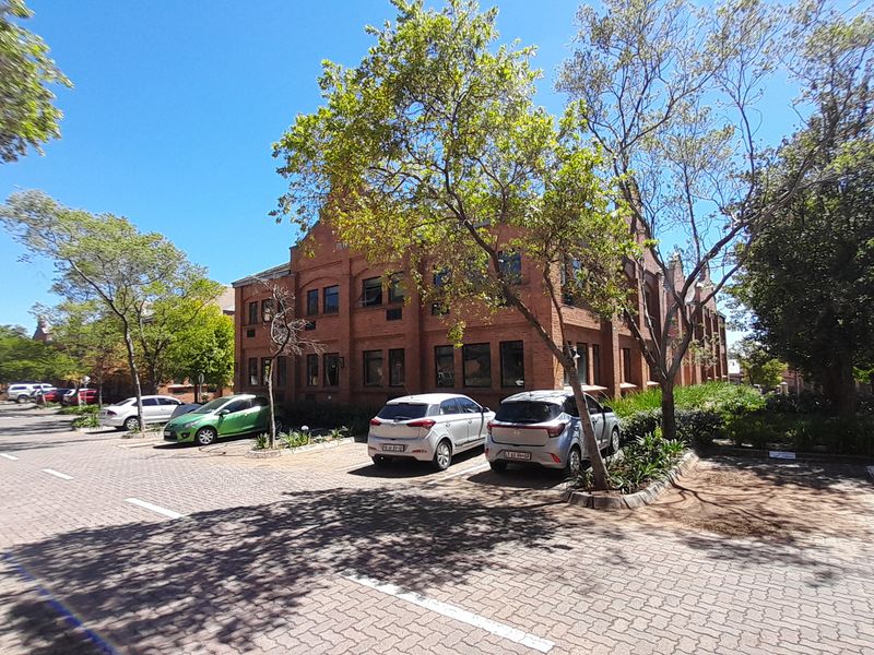 A view of the front exterior of a brick office building with an adjacent paved parking area and mature trees.