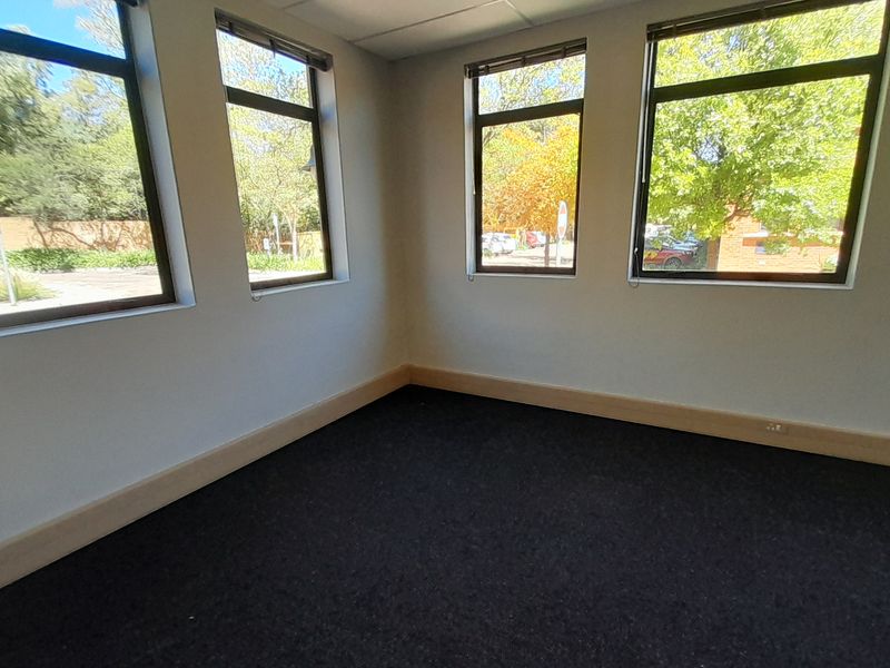 A bright, empty office space featuring large windows providing natural light, dark carpeting, and neutral wall finishes, suitable for a professional workspace.