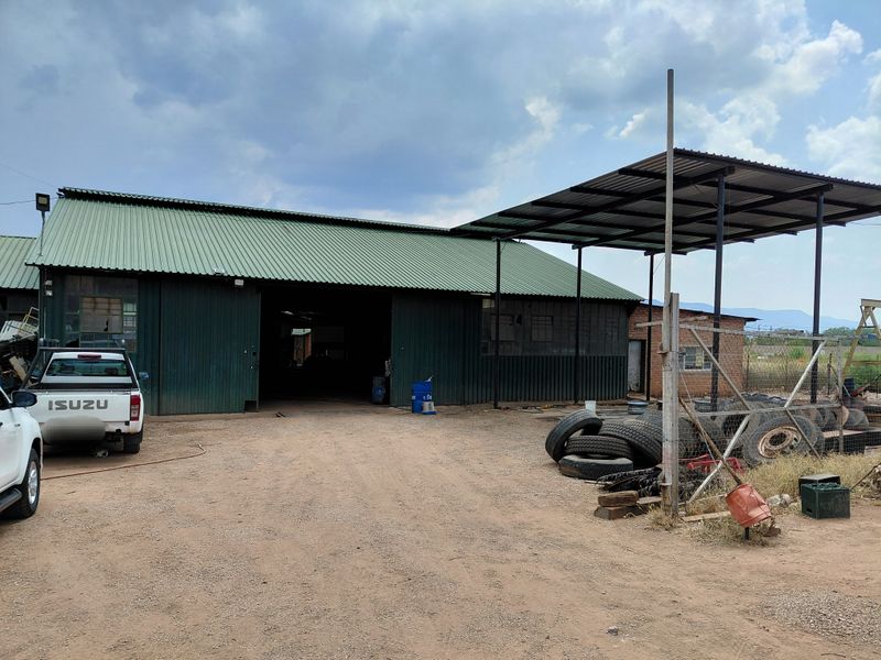 Industrial warehouse facility featuring green metal cladding, a large open entrance, and an attached covered outdoor work area on a gravel yard.