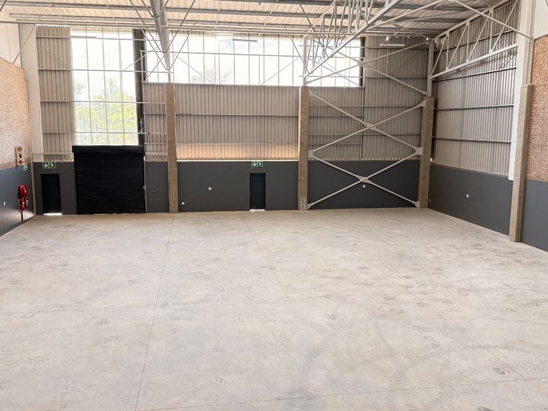 Spacious industrial warehouse unit featuring high ceilings with exposed steel trusses, concrete flooring, a large roller shutter door, and integrated fire safety systems.