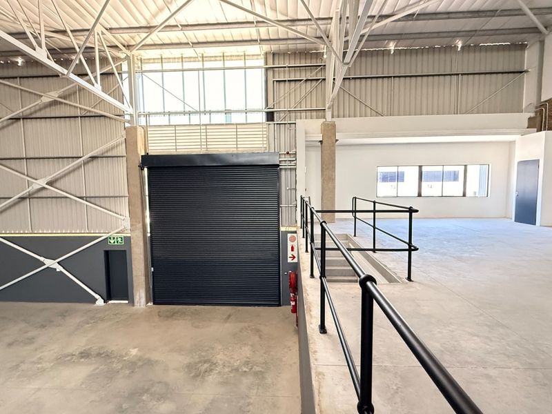 Spacious industrial warehouse unit featuring high ceilings with exposed steel trusses, concrete flooring, a large roller shutter door, and integrated safety features.