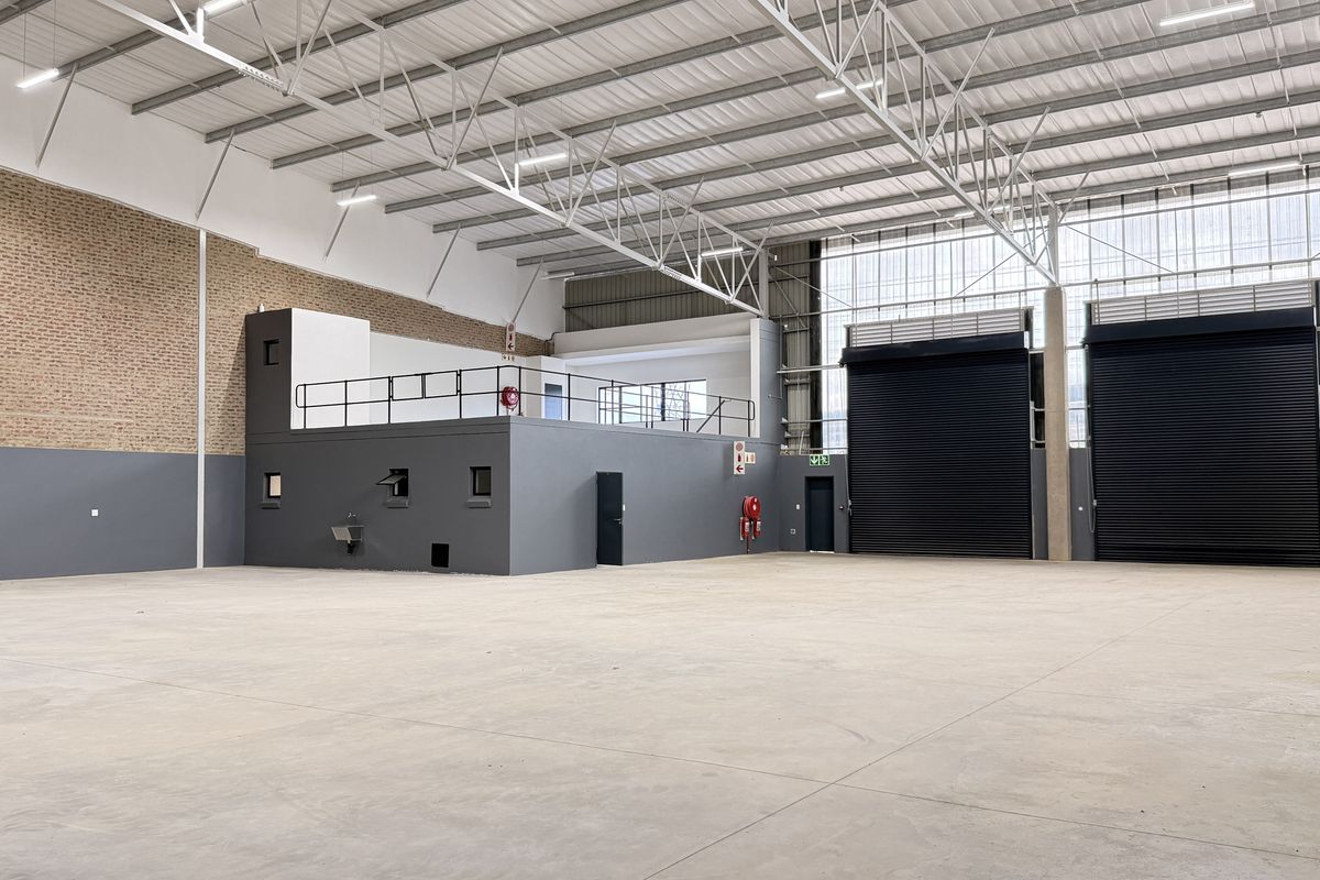 Spacious industrial warehouse featuring high ceilings with exposed steel trusses, concrete flooring, dual roller shutter doors, and a functional mezzanine level.