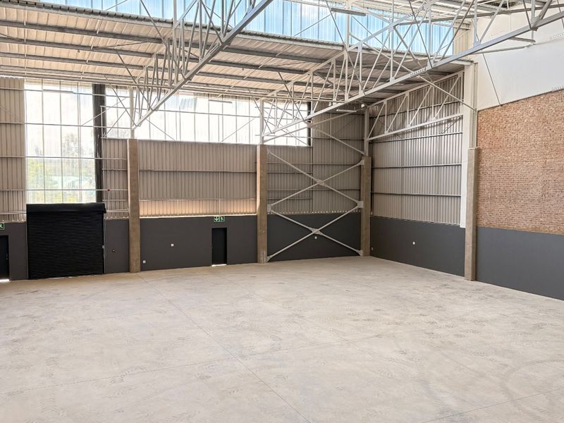 Spacious industrial warehouse unit featuring high ceilings with exposed steel trusses, concrete flooring, and a large roller shutter door for easy access.