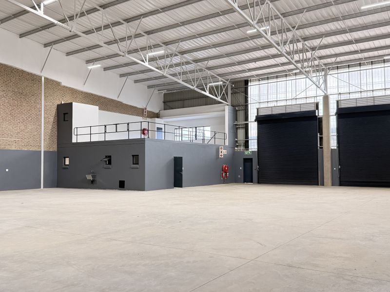 Spacious industrial warehouse featuring high ceilings with exposed steel trusses, concrete flooring, dual roller shutter doors, and a functional mezzanine level.