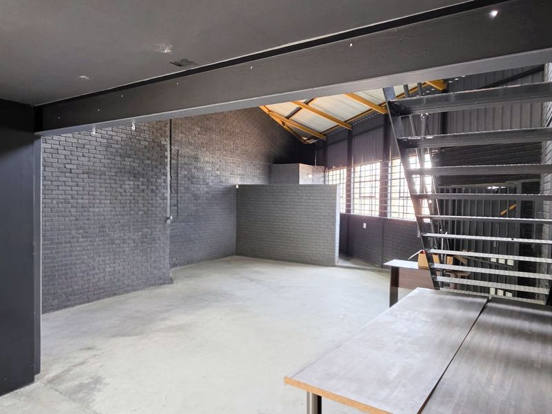 Spacious industrial open-plan area featuring exposed brick walls, concrete flooring, steel structural beams, and a metal staircase leading to an upper level.