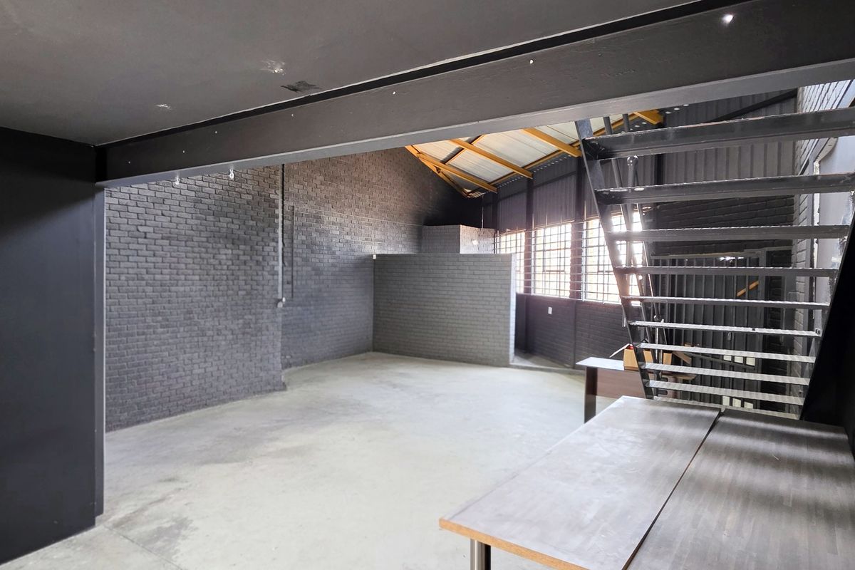 Spacious industrial open-plan area featuring exposed brick walls, concrete flooring, steel structural beams, and a metal staircase leading to an upper level.