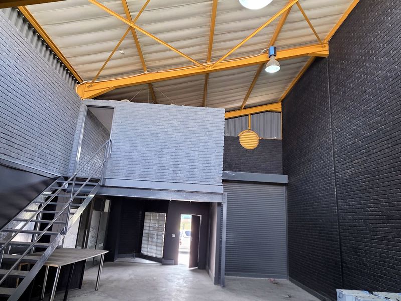 Spacious industrial unit featuring high ceilings with exposed steel trusses, a mezzanine level, metal roller shutter door, and modern dark-painted brick walls.