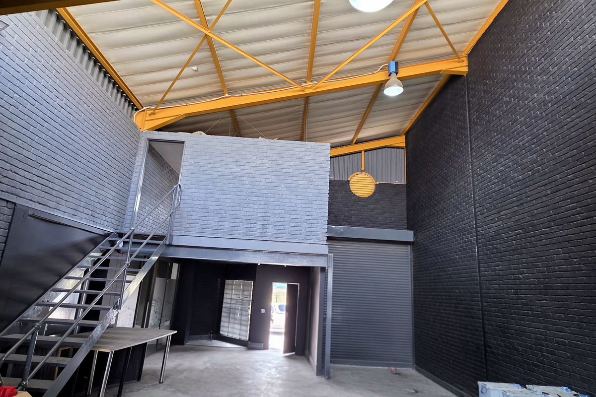 Spacious industrial unit featuring high ceilings with exposed steel trusses, a mezzanine level, metal roller shutter door, and modern dark-painted brick walls.