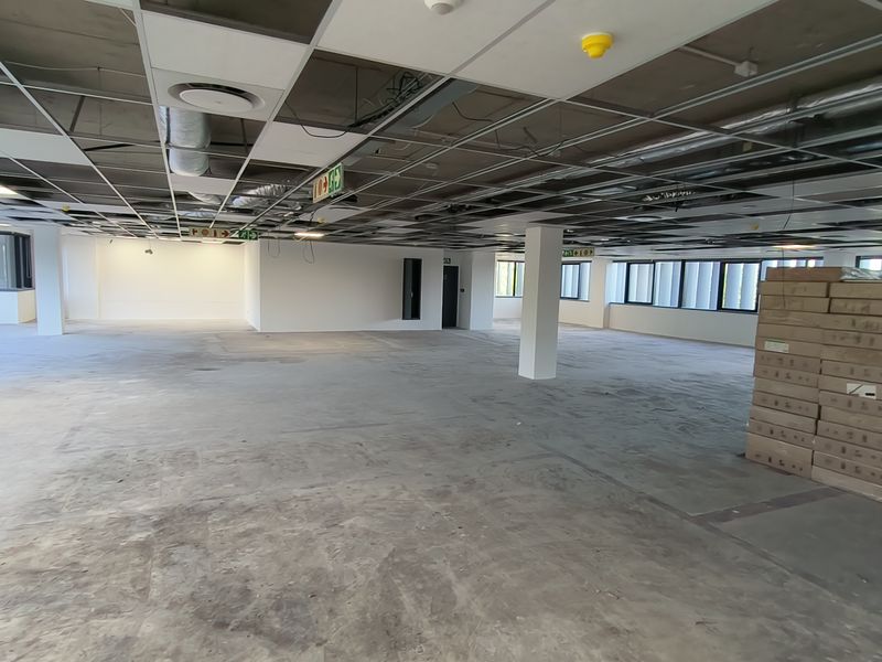 A large, open-plan office space currently in a shell condition with exposed ceiling infrastructure, concrete flooring, and large windows providing natural light.